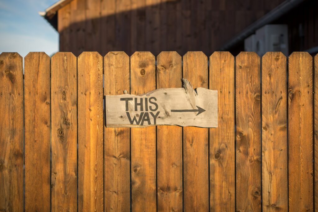 Ein Holzzaun, an dem ein altes Holzschild hängt. Darauf ein Pfeil nach rechts und die Beschriftung "This way". 