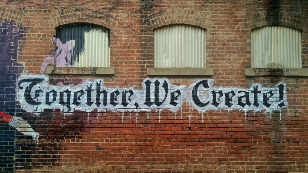 Ausschnitt eines alten Gebäudes mit Ziegelmauer und abgedeckten Fenstern. Darauf als Graffiti gesprüht ein Pinsel und der Spruch "Together we create".