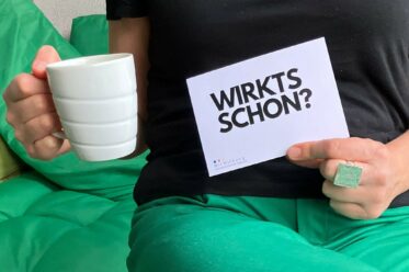 Eine Person, in einer Hand eine Kaffeetasse, in der anderen ein Schild mit der aufschrift "Wirt's schon?"