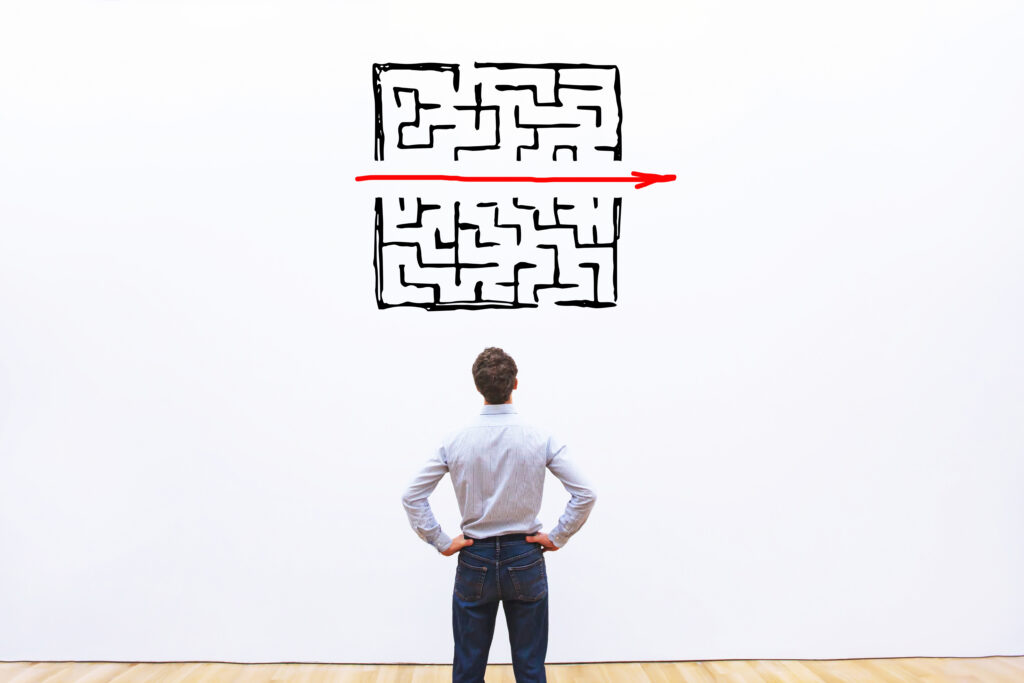 Ein Mann steht vor einer Wand. Auf die Wand ist ein Labyrinth gemalt, das in der Mitte geteilt ist. Ein roter Pfeil weist durch die Schneise und zeigt so einen einfachen Weg durch das Labyrinth auf.