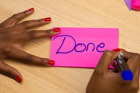 Zwei Hände mit rot lackierten Fingernägeln, die einen pinken Klebezettel halten und mit "Done" beschriften. 