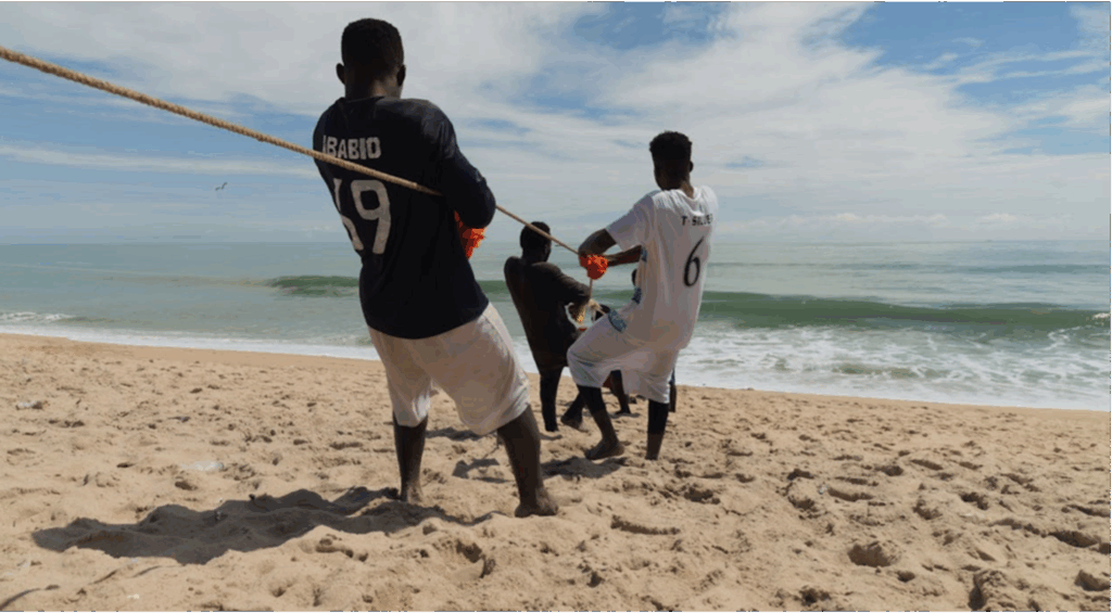 Mehrere Männer stehen am Strand am Meer und ziehen gemeinsam an einem Seil - vermutllich, um, ein Boot aus dem Waser zu ziehen. 