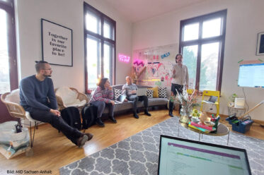 Projekttreffen im Zuge der ebenenübergreifenden Projektarbeit. Zu sehen sind Tobias Krüger und drei weitere Personen in einer kreativen Arbeitsumgebung. Bunte Möbel, buntes Arbeitsmaterial. Im Vordergrund ist ein Laptopbildschirm zu sehen, im Hintergrund ein großer Bildschirm, auf dem Inhalte präsentiert werden.