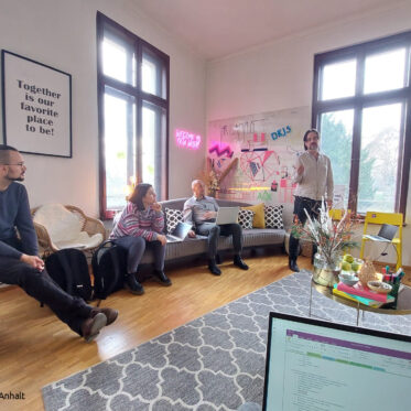 Projekttreffen im Zuge der ebenenübergreifenden Projektarbeit. Zu sehen sind Tobias Krüger und drei weitere Personen in einer kreativen Arbeitsumgebung. Bunte Möbel, buntes Arbeitsmaterial. Im Vordergrund ist ein Laptopbildschirm zu sehen, im Hintergrund ein großer Bildschirm, auf dem Inhalte präsentiert werden.