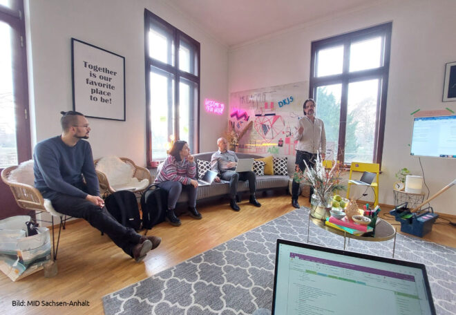 Projekttreffen im Zuge der ebenenübergreifenden Projektarbeit. Zu sehen sind Tobias Krüger und drei weitere Personen in einer kreativen Arbeitsumgebung. Bunte Möbel, buntes Arbeitsmaterial. Im Vordergrund ist ein Laptopbildschirm zu sehen, im Hintergrund ein großer Bildschirm, auf dem Inhalte präsentiert werden.