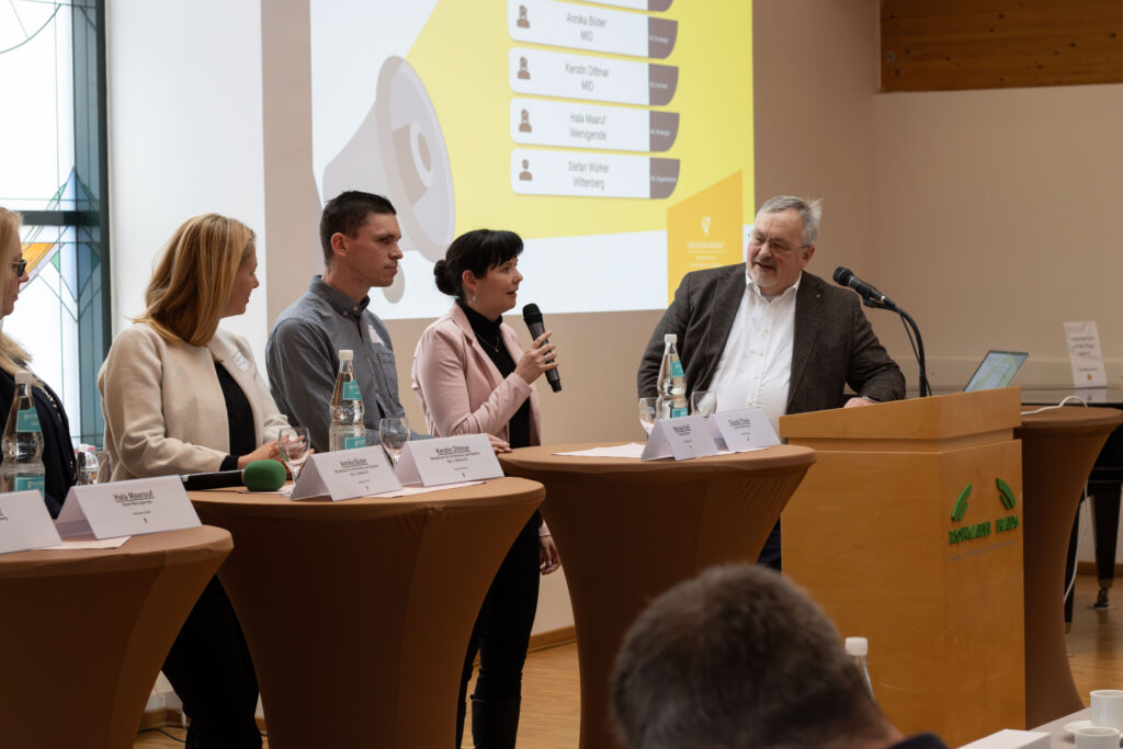 Projekttreffen zur Halbzeit des GDST-Projektes. Auf der Bühne mehrere Vertreter:innen von Kommunen und ein Moderator. Dahinter eine Leinwand auf der eine Folie mit den Namen der Personen zu sehen ist. 