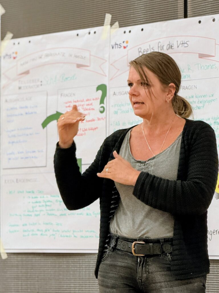 Eine Mitarbeiterin der VHS steht vorn beim Hackathon vor einem Flipchart „Reels für die VHS“ und präsentiert Ergebnisse. Dabei gestikuliert sie mit den Händen.