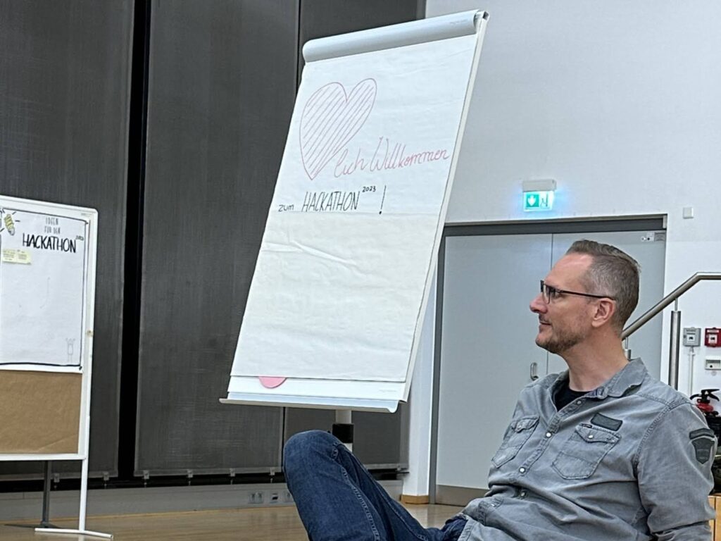 Stephan Rinke sitzt lässig auf einem Stuhl und schaut nach vorn. Neben ihm ist das Willkommens-Flipchart zu sehen. 