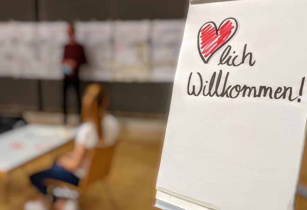 Foto einer Szene vom Hackathon. Im Vordergrund ein Flipchart mit „Herzlich Willkommen!“. Im hintergrund verschwommen eine Person die vor einer Wand mit einer Reihe von Flipcharts steht. An einem Tisch sitzt eine Person und schaut nach vorn.