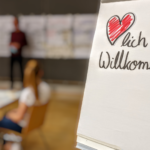 Foto einer Szene vom Hackathon. Im Vordergrund ein Flipchart mit „Herzlich Willkommen!“. Im hintergrund verschwommen eine Person die vor einer Wand mit einer Reihe von Flipcharts steht. An einem Tisch sitzt eine Person und schaut nach vorn.