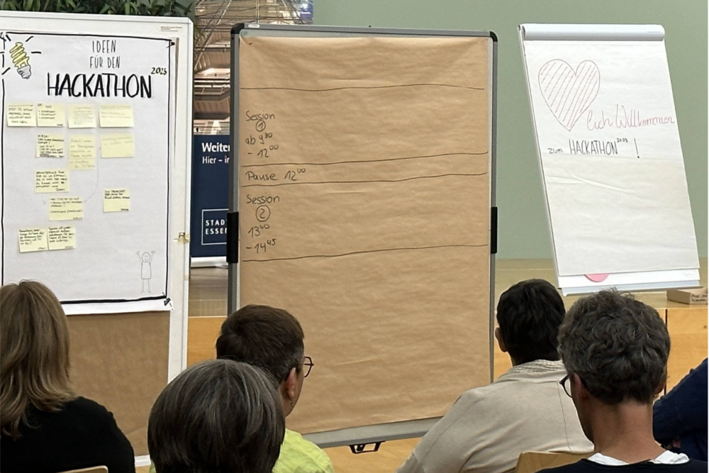 Szene zu Beginn des Hackathons. Menschen von hinten fotografiert im sitzen im Plenum. Vorn stehen Pinnwände für die Themenplanung und ein Willkommens-Flipchart.
