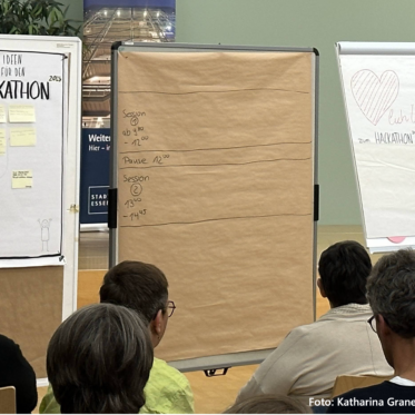 Szene zu Beginn des Hackathons. Menschen von hinten fotografiert im sitzen im Plenum. Vorn stehen Pinnwände für die Themenplanung und ein Willkommens-Flipchart.
